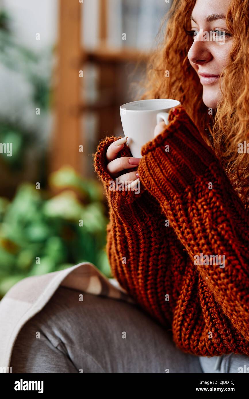 A cute ginger girl with curly hair is sitting in the chair at home in the morning and drinking her coffee. Stock Photo