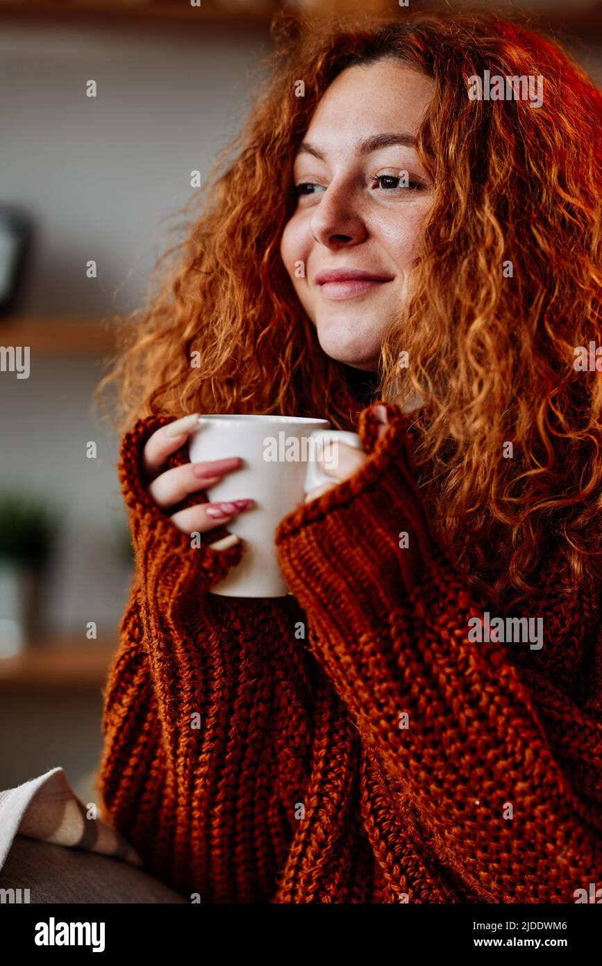 A cute young woman with red curly hair is enjoying her cup of coffee in the morning. Stock Photo