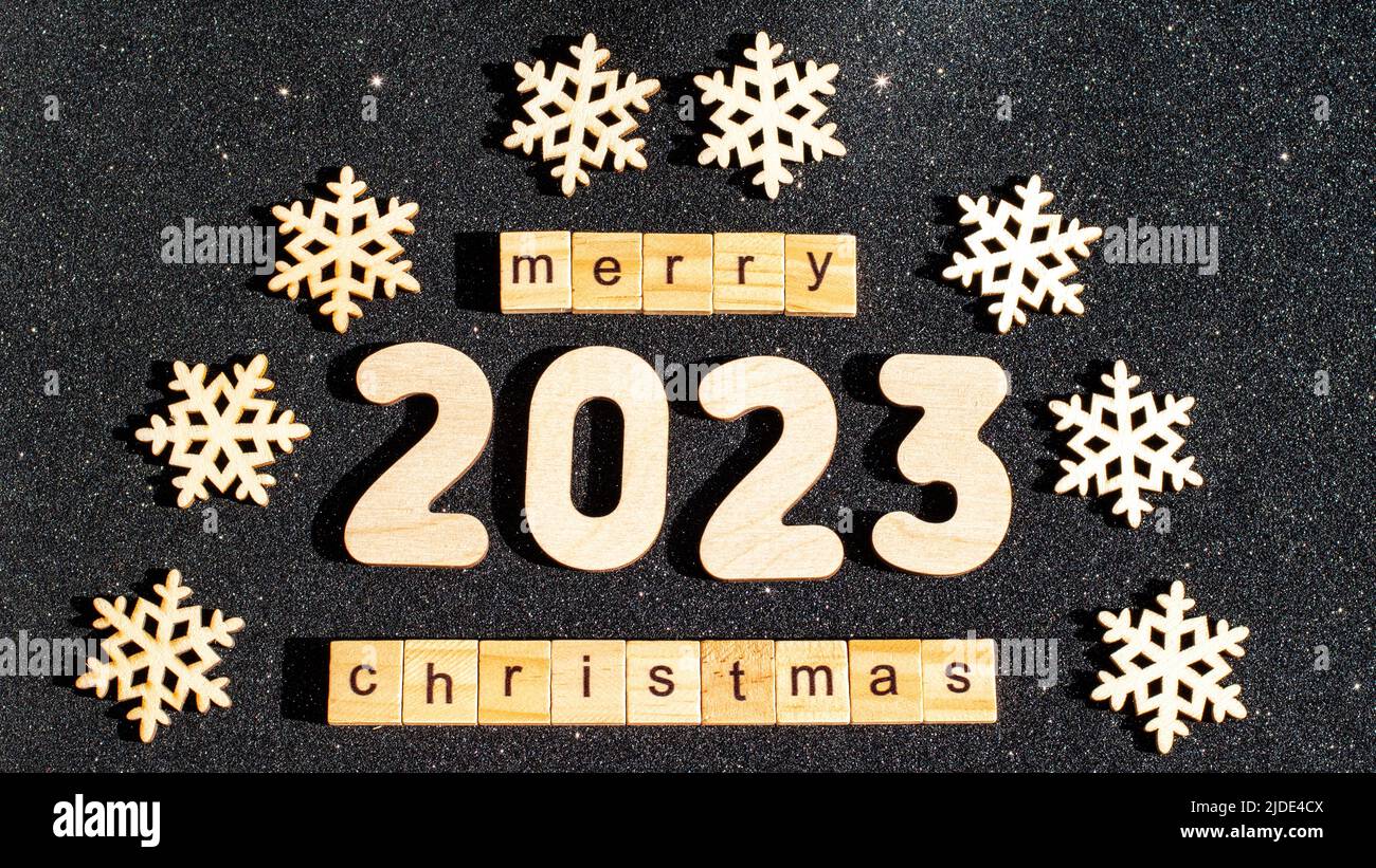 Concept for Christmas and New Year - Wooden snowflakes numbers 2023 and the inscription HAPPY CHRISTMAS from wooden letters on black glittering starry Stock Photo