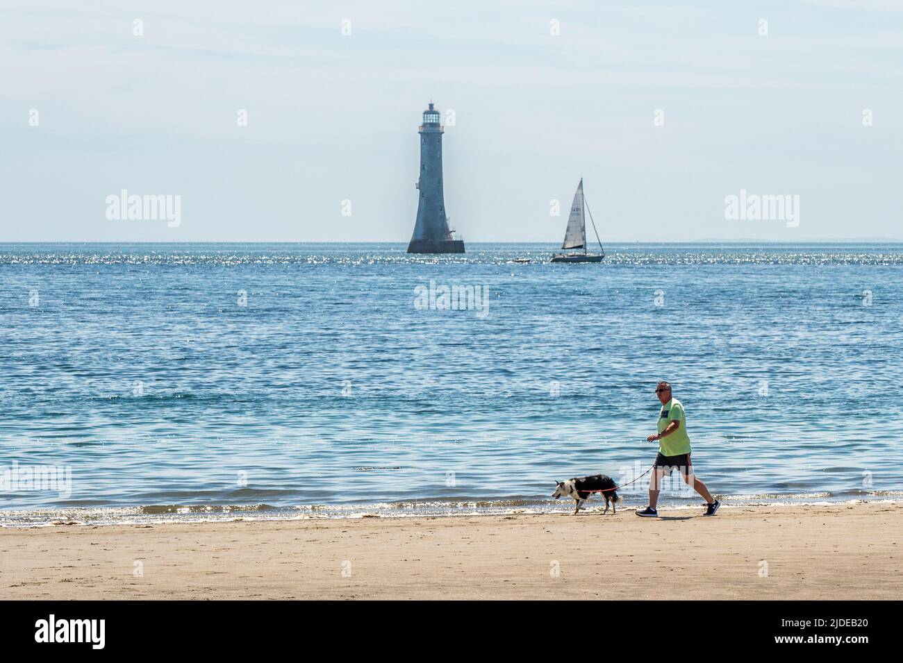 Cranfield, County Down, UK. On a day of sunshine and highs of 20C, many people hit the beaches in Northern Ireland. A man walks his dog along the shoreline of Cranfield Beach with the Haulbowline Lighthouse as a backdrop. Credit: AG News/Alamy Live News Stock Photo