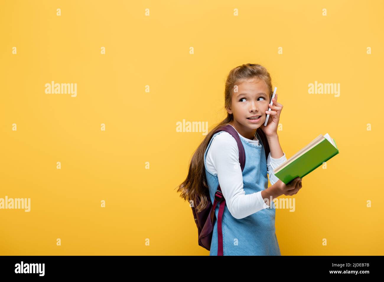 Schoolgirl talking on smartphone and holding book isolated on yellow Stock Photo