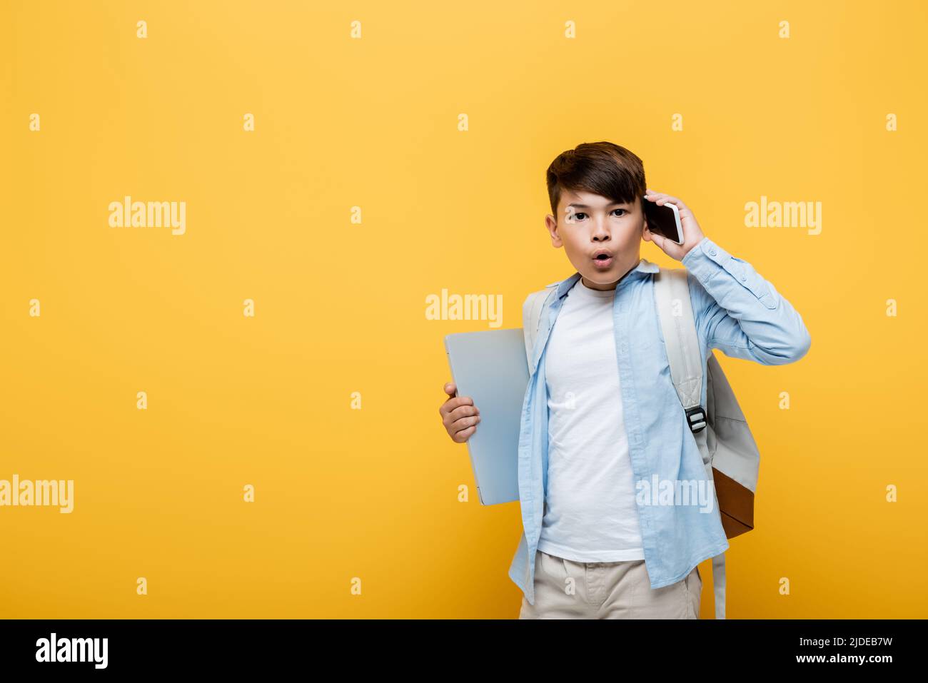 Shocked asian schoolboy talking on smartphone and holding laptop isolated on yellow Stock Photo