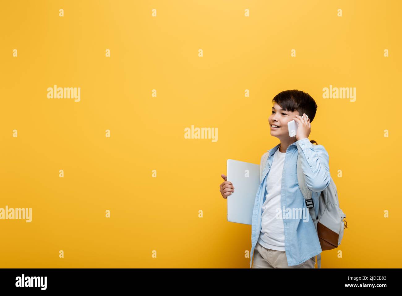 Smiling asian schoolboy talking on smartphone and holding laptop isolated on yellow Stock Photo