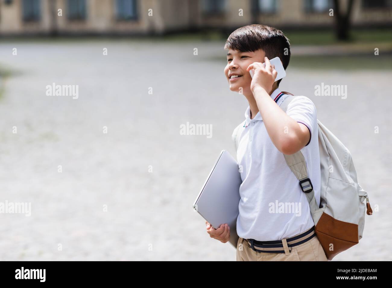 Smiling asian schoolboy holding laptop and talking on mobile phone outdoors Stock Photo