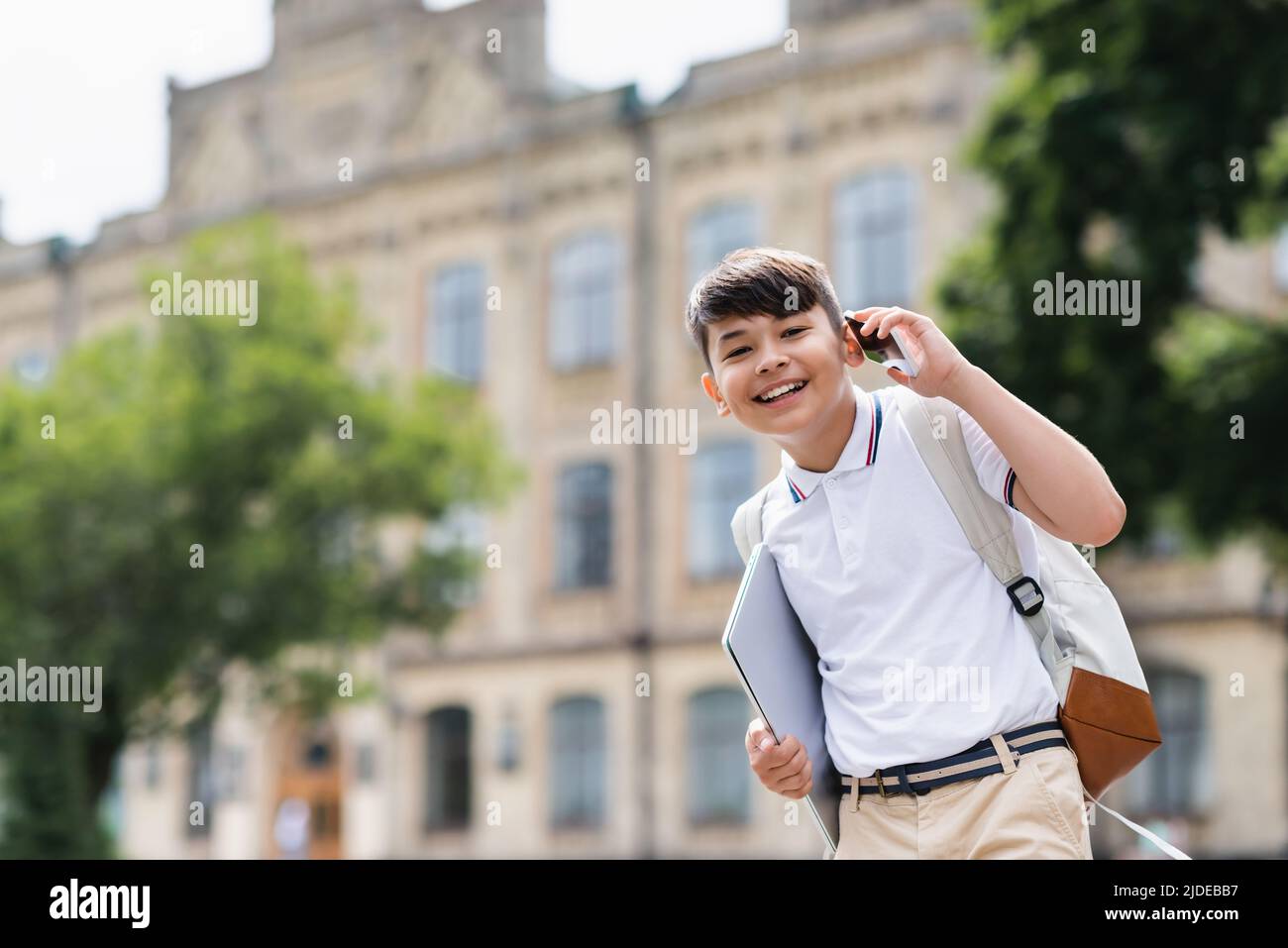Cheerful asian schoolboy holding laptop and talking on smartphone outdoors Stock Photo