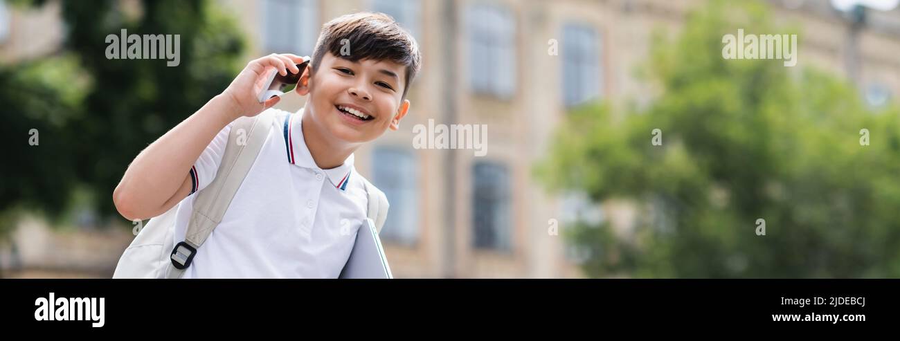 Asian schoolboy talking on cellphone and holding laptop outdoors, banner Stock Photo