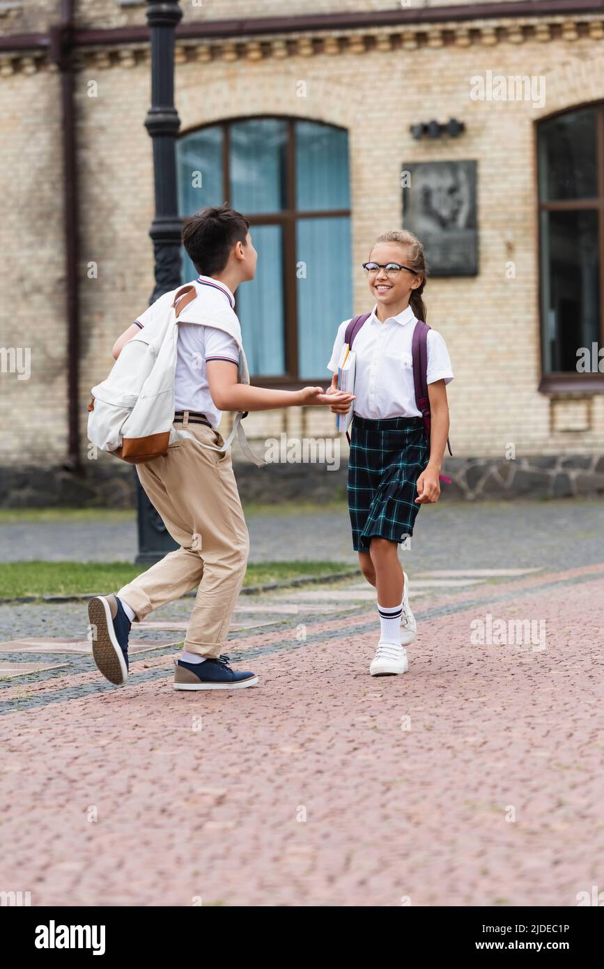 Asian schoolboy with backpack talking to cheerful friend with notebooks ...