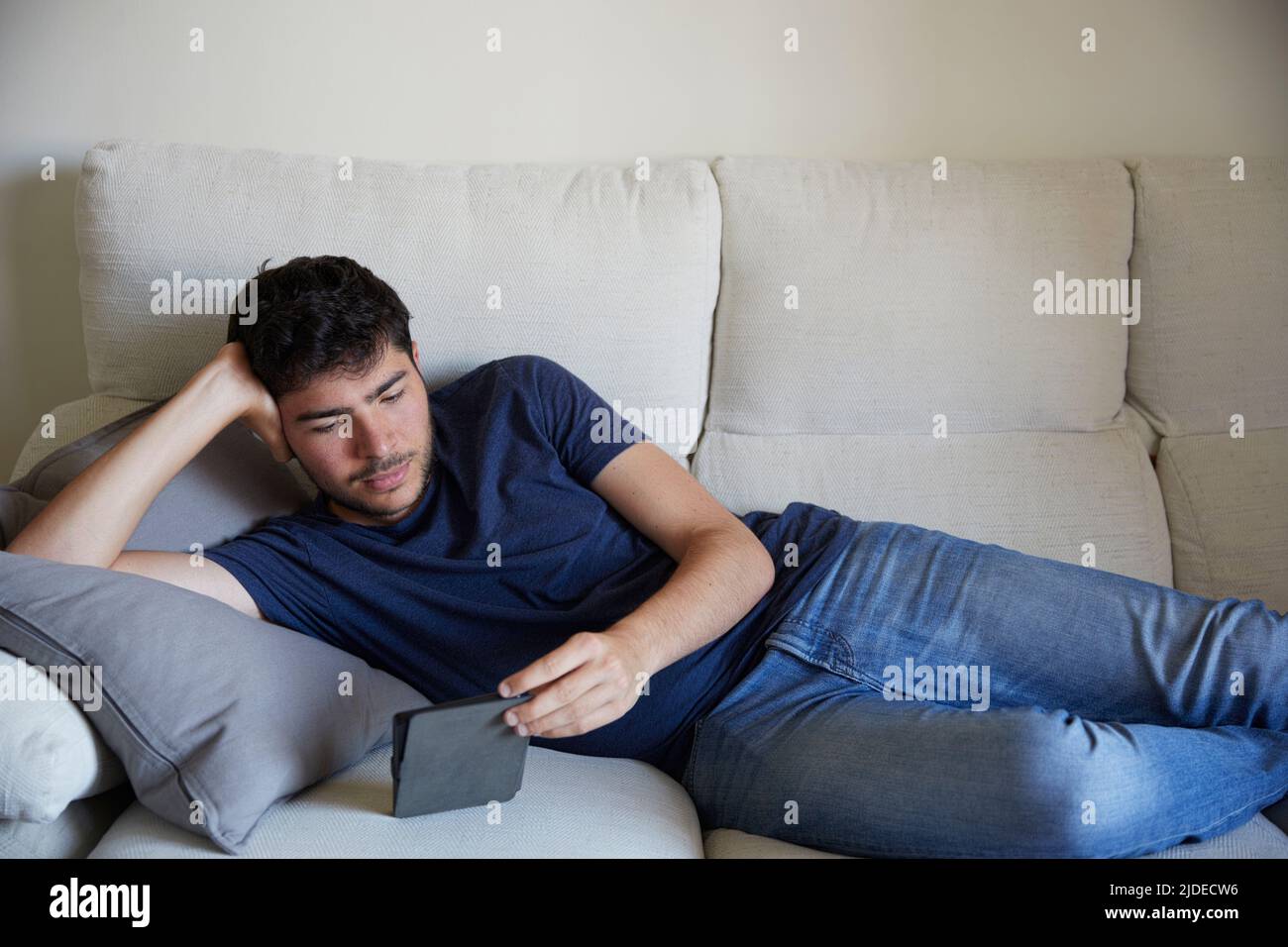 Young man lying down on the sofa reading an ebook Stock Photo