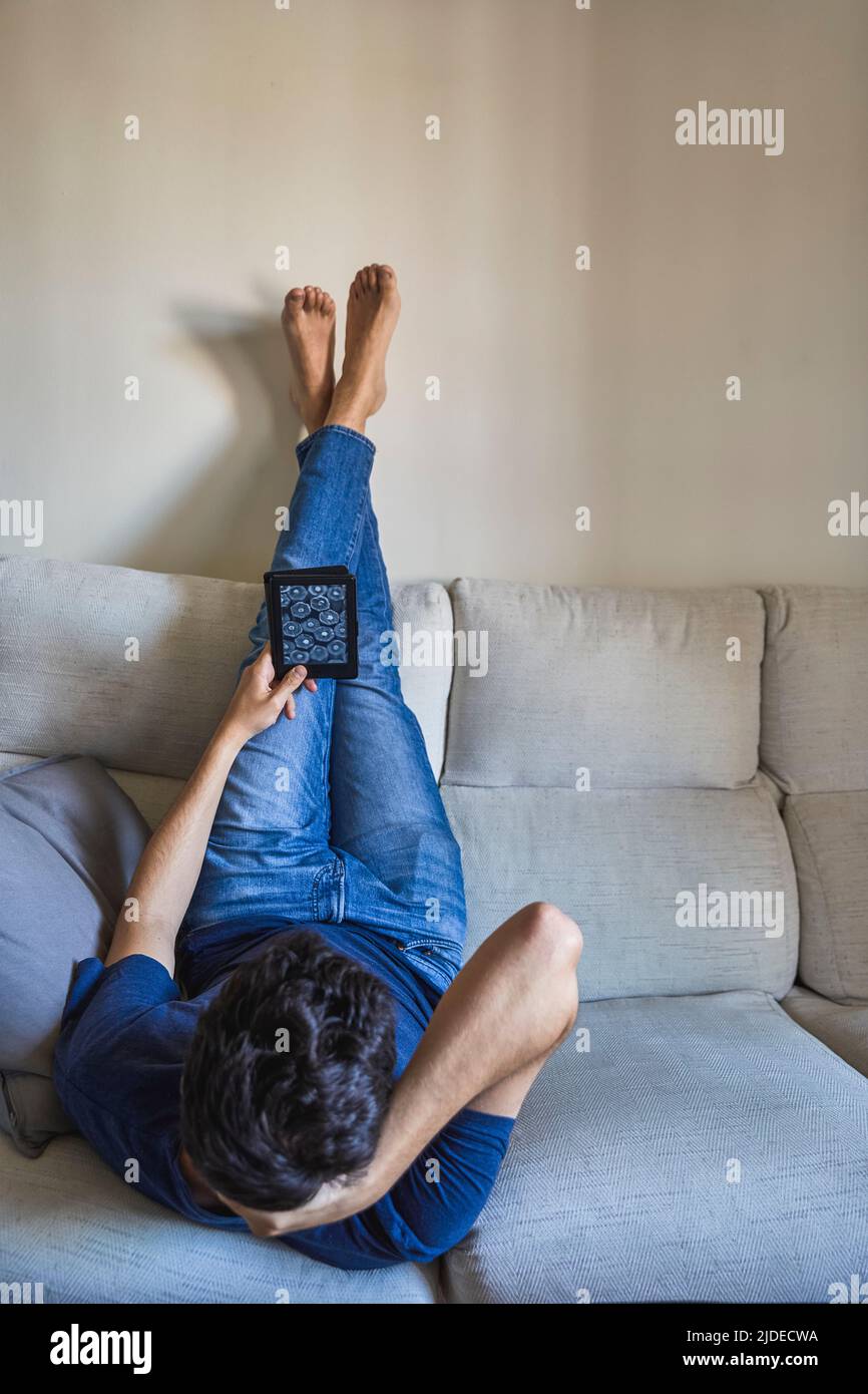 Young man lying with his feet on the wall face down reading an ebook on the sofa Stock Photo