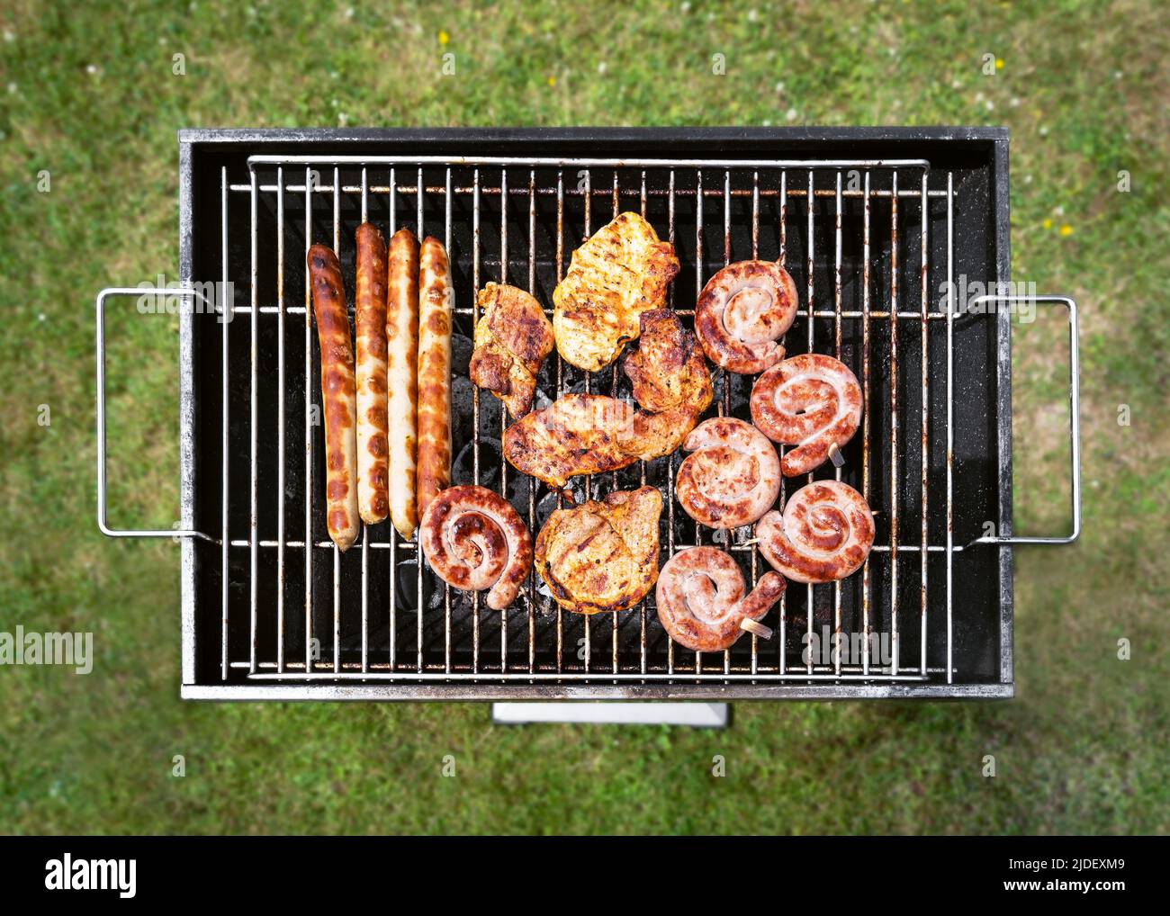 Tasty assortment of meat on a summer barbecue with steaks, chicken, sausages, spirals beef sausage. Overhead view over green grass. Stock Photo