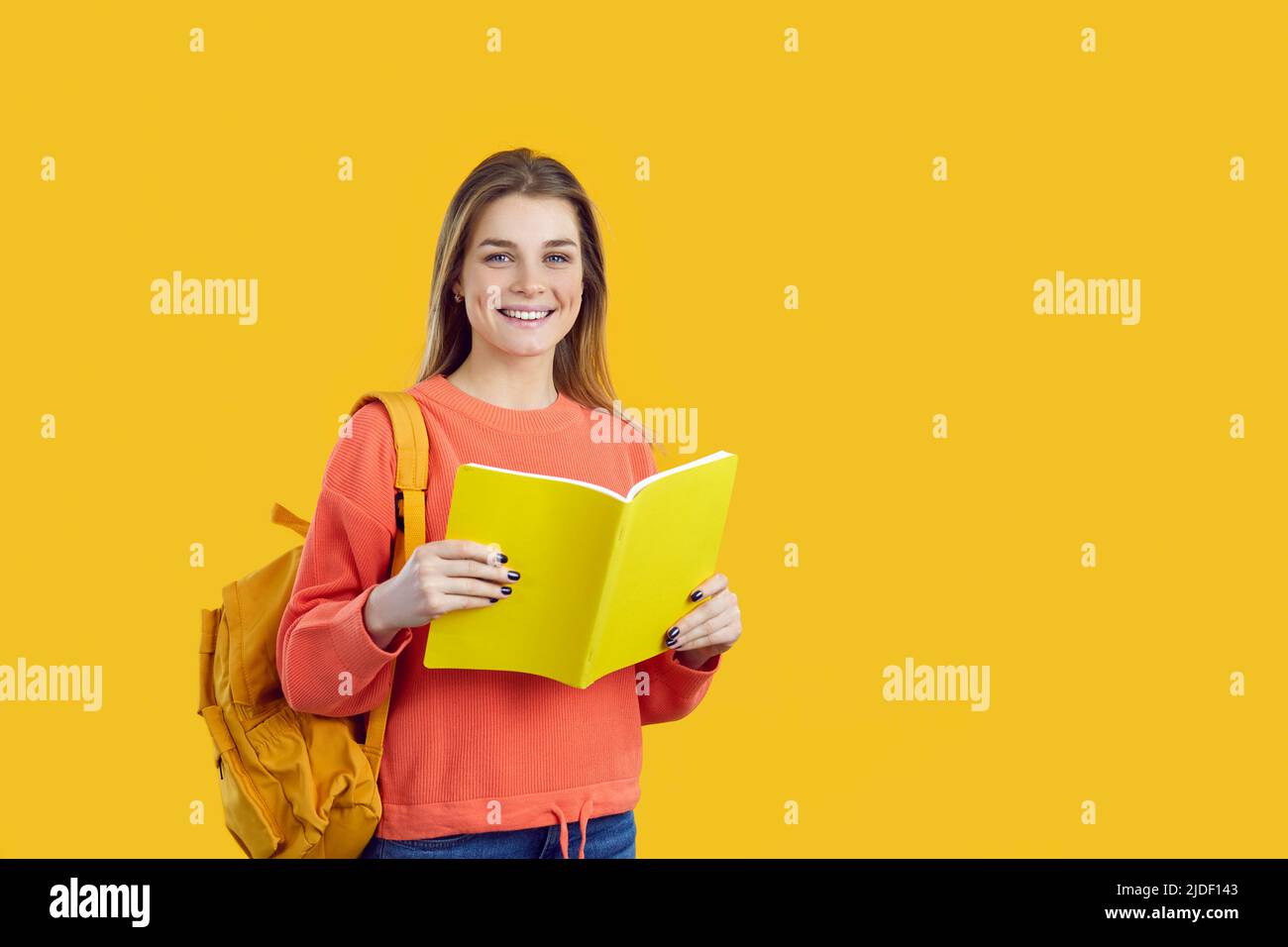 Portrait of smiling female student with backpack and textbook isolated on yellow background. Stock Photo