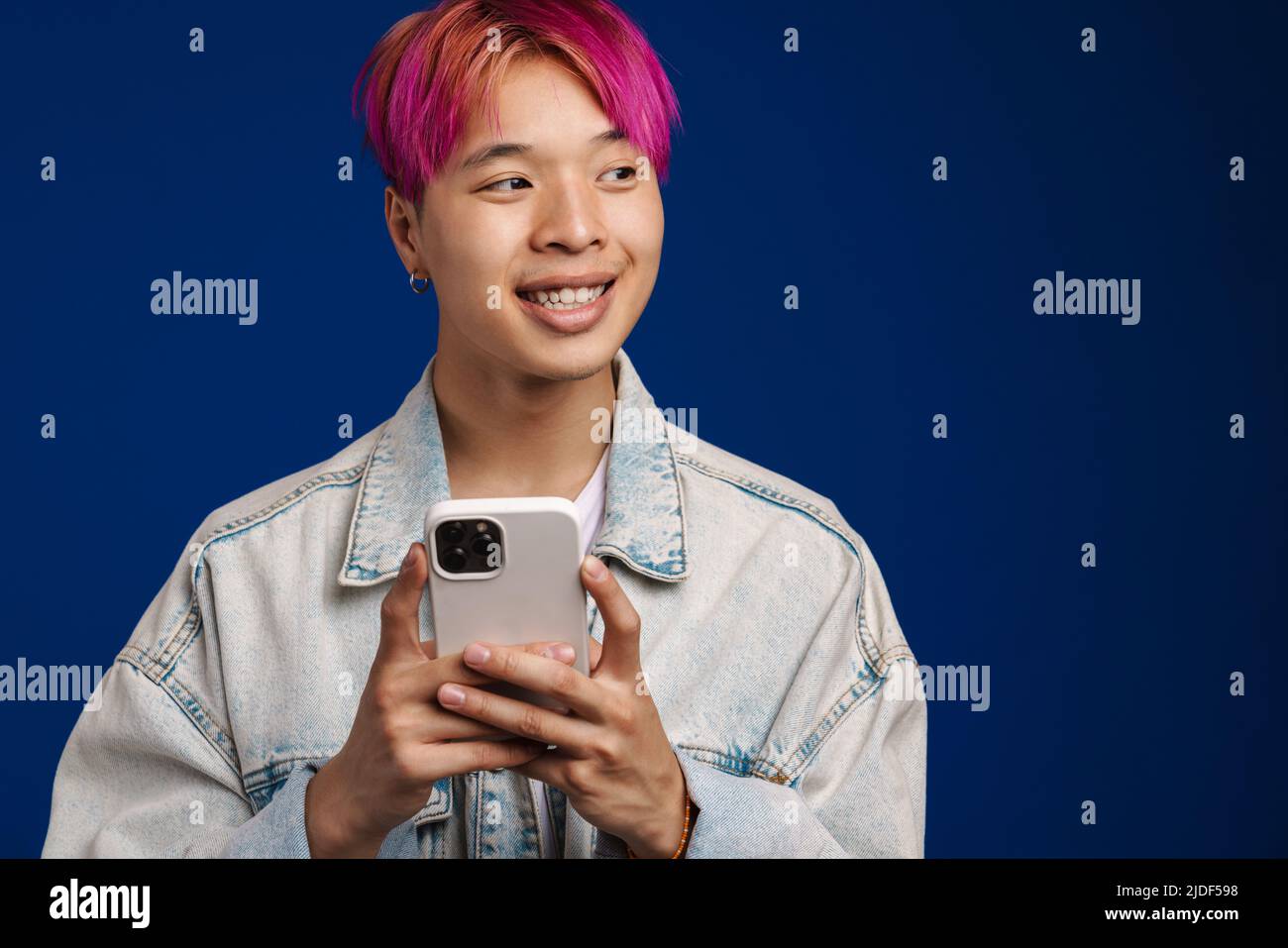 Asian boy in denim jacket smiling while using cellphone isolated over blue background Stock Photo