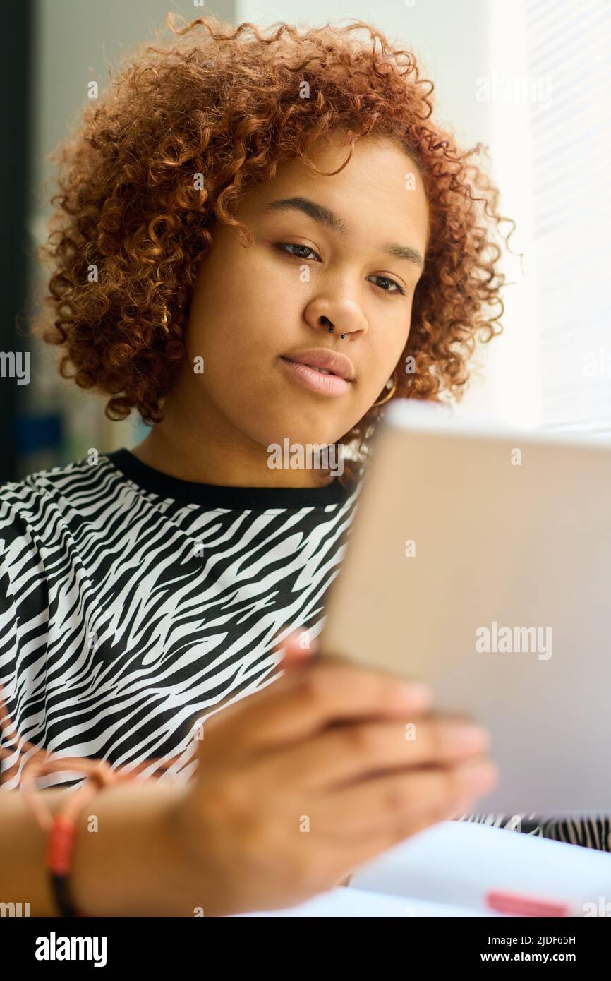 African American girl looking through online table or electronic manual while holding digital tablet in front of her face at lesson Stock Photo