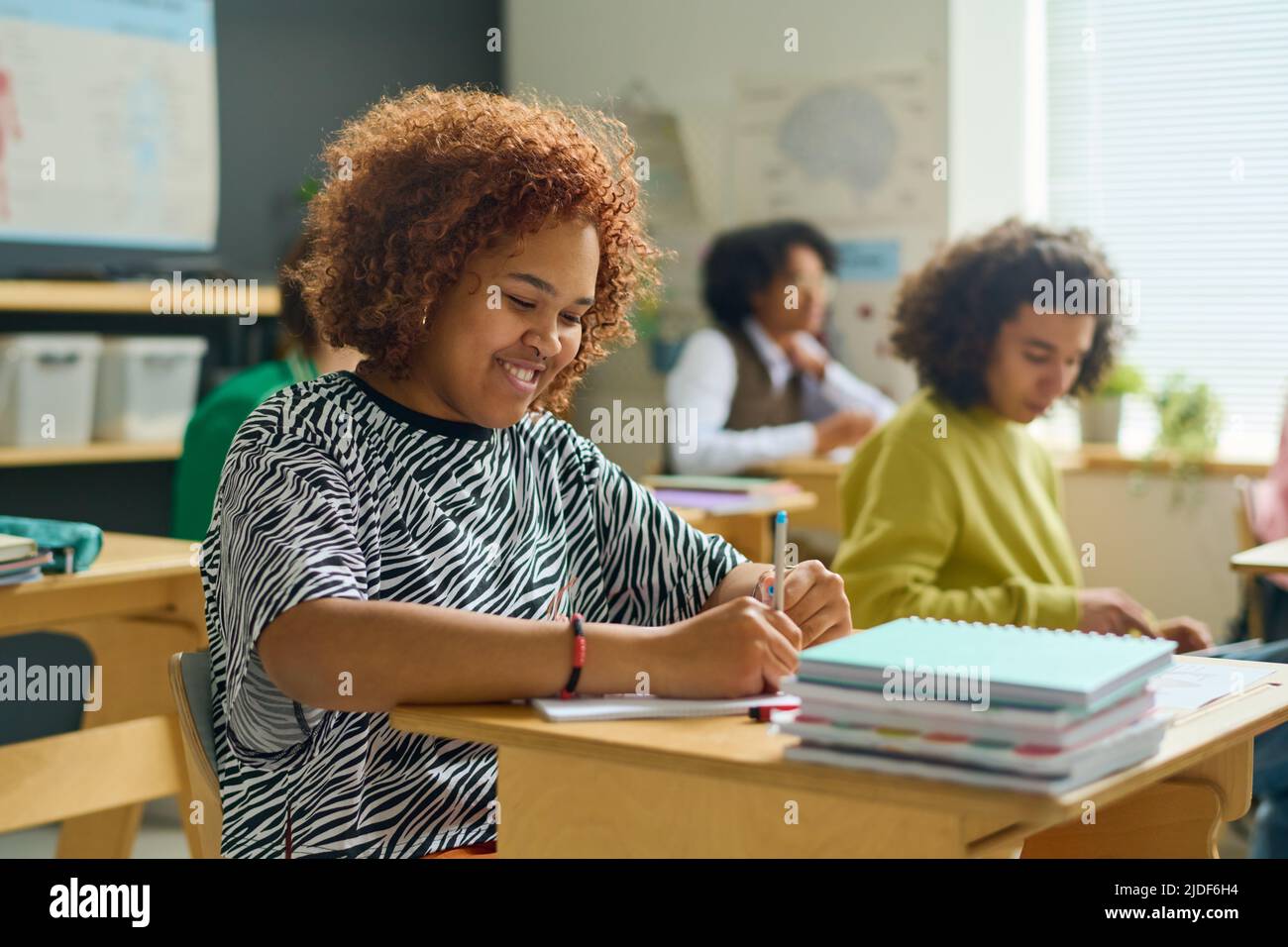 Happy female teenager with pen making notes in copybook during lecture or individual assignment against classmates at lesson Stock Photo