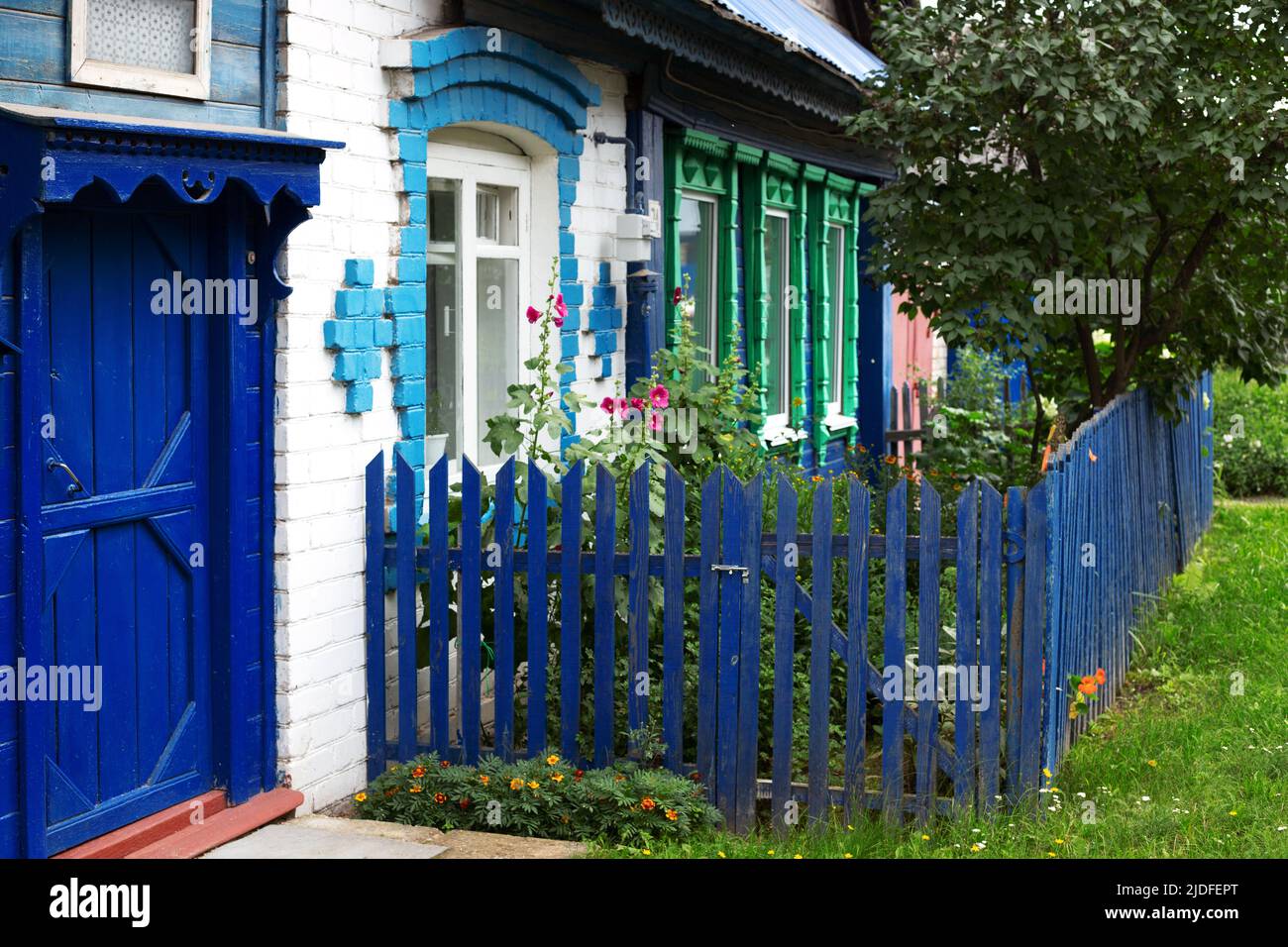 Old wooden and white brick house with green carved window frames, blue door and front garden fence Stock Photo