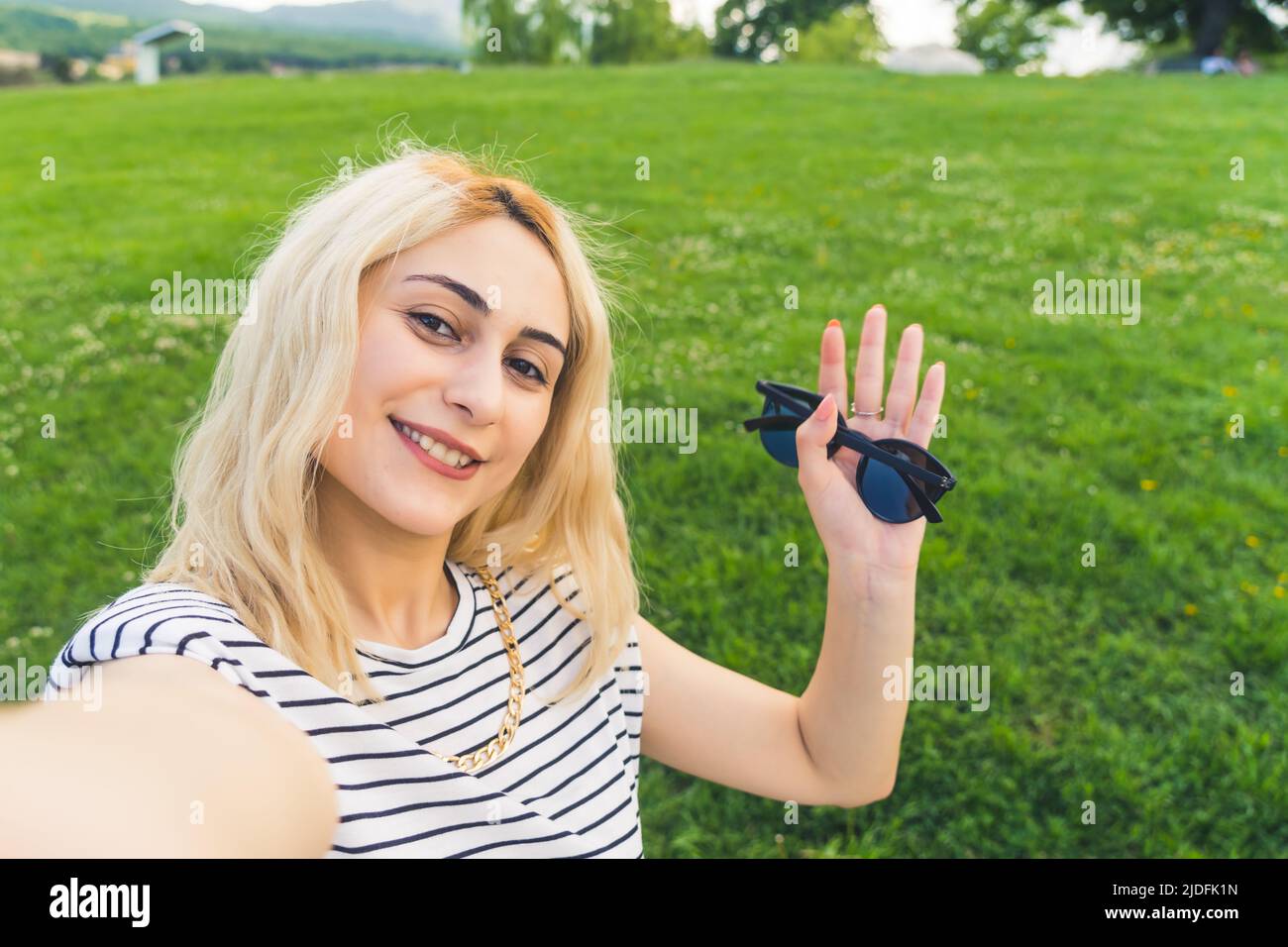 young blond Caucasian girl taking a selfie and waving with a sunglasses in a hand. medium closeup park . High quality photo Stock Photo
