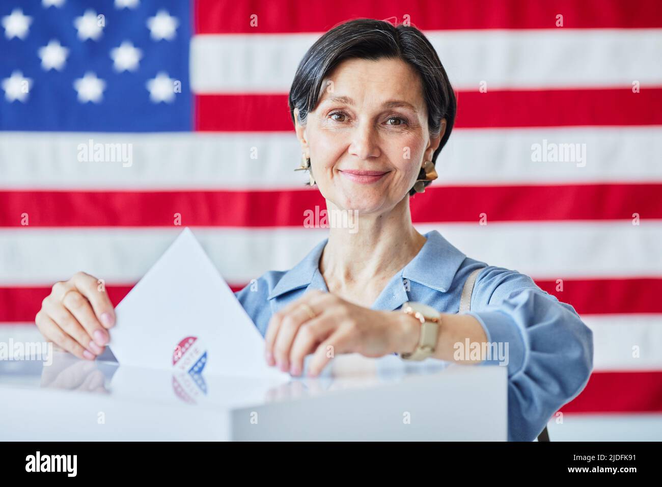 Portrait of smiling adult woman putting ballot in bin against American