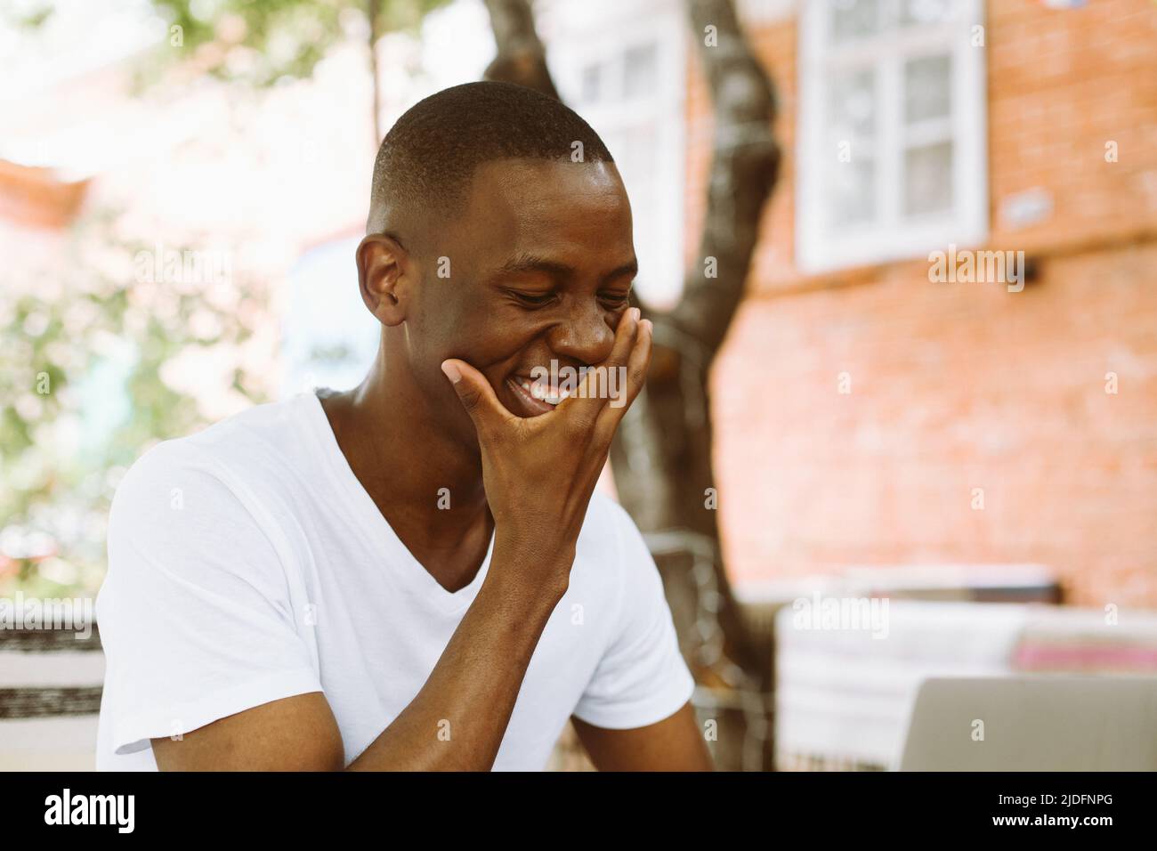 Smiling, laughing african american businessman working on laptop, covering his face by hand in the street cafe outdoors. Stock Photo
