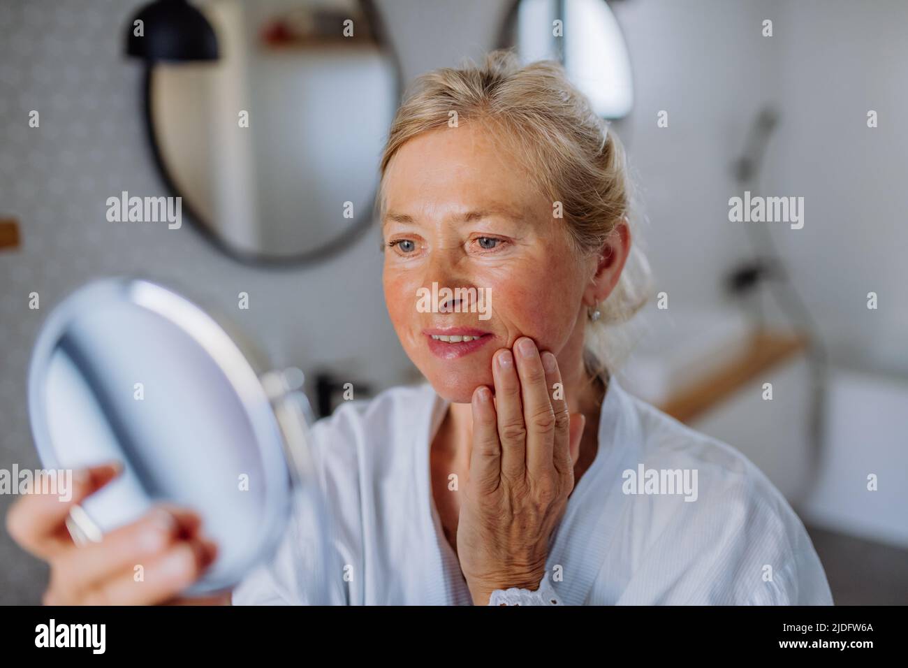 Beautiful senior woman in bathrobe looking at mirror and applying natural face cream in bathroom, skin care concept. Stock Photo