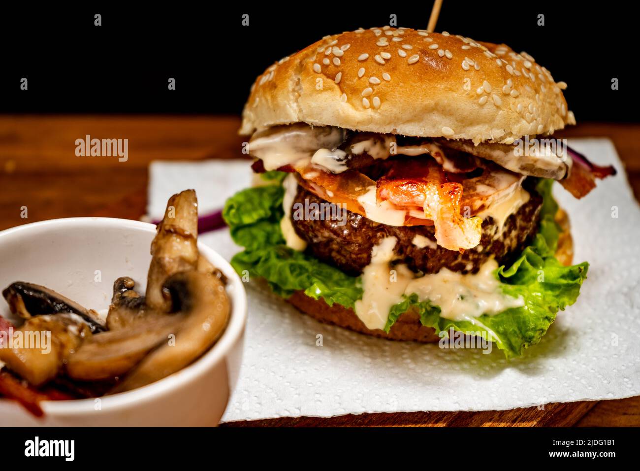 Homemade beef burger with lettuce, tomato, mushrooms, bacon and cheese on a wooden board with black background. Chopped view. Copy text. Home made, fa Stock Photo