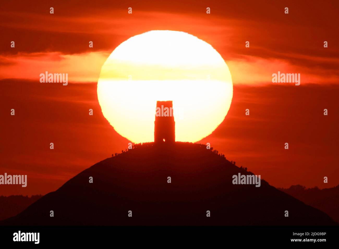 Glastonbury, Somerset, UK.  21st June 2022.  UK Weather:  The sky glows orange as the sun rises up from behind St Michael’s Tower on Glastonbury Tor in Somerset which is packed with revellers at sunrise during the summer solstice.  Picture Credit: Graham Hunt/Alamy Live News Stock Photo