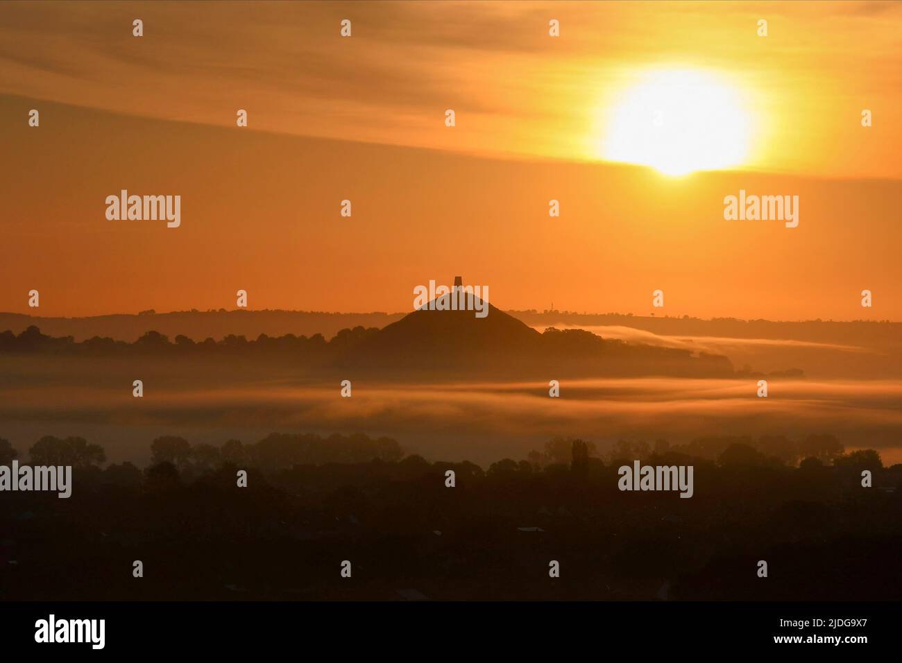 Glastonbury, Somerset, UK.  21st June 2022.  UK Weather:  The sky glows orange as the sun rises up from above St Michael’s Tower on Glastonbury Tor in Somerset which is packed with revellers at sunrise during the summer solstice.  Picture Credit: Graham Hunt/Alamy Live News Stock Photo