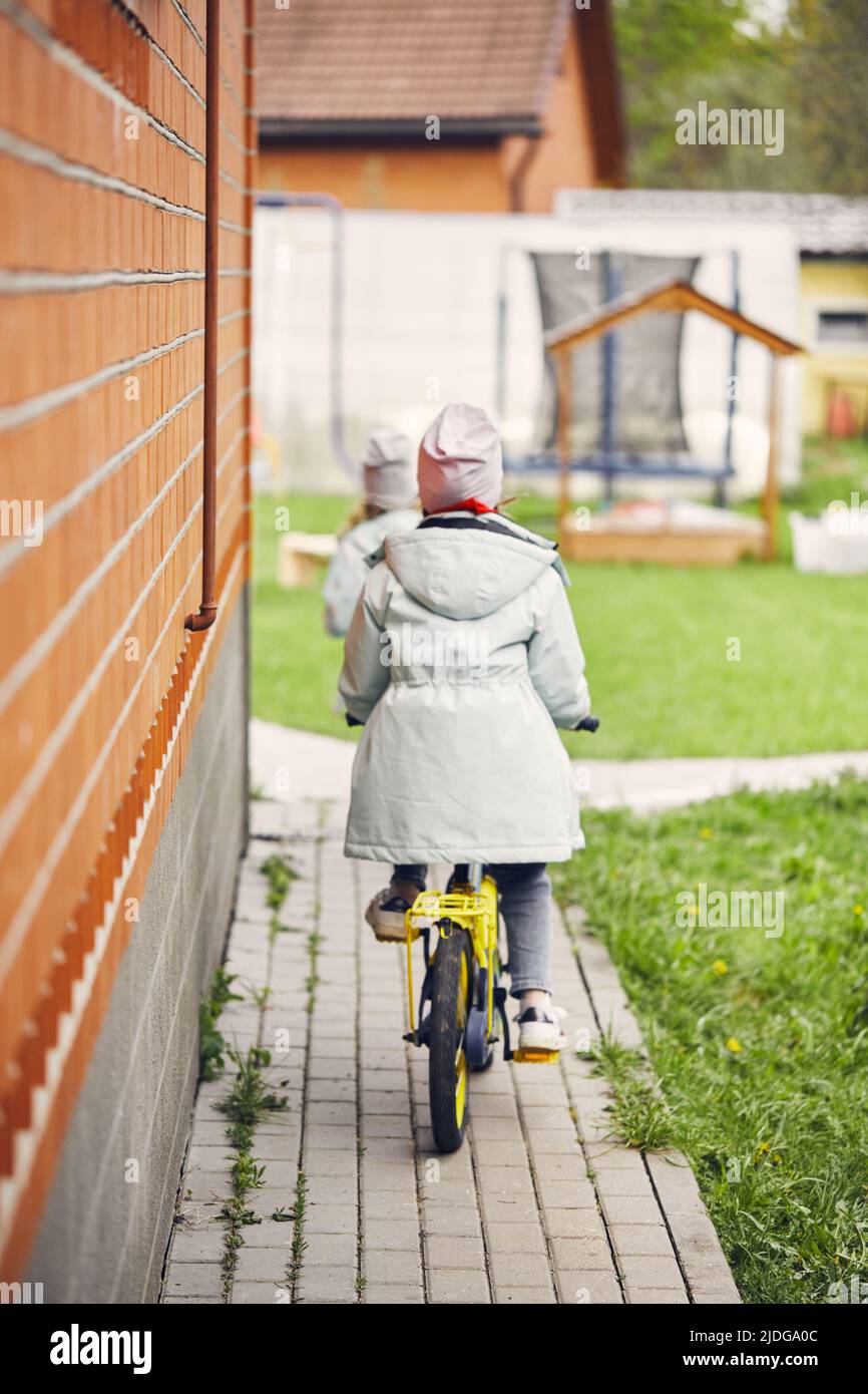 Little girls on bicycles ride around the yard. The concept of a happy childhood. Vertical photo. Stock Photo