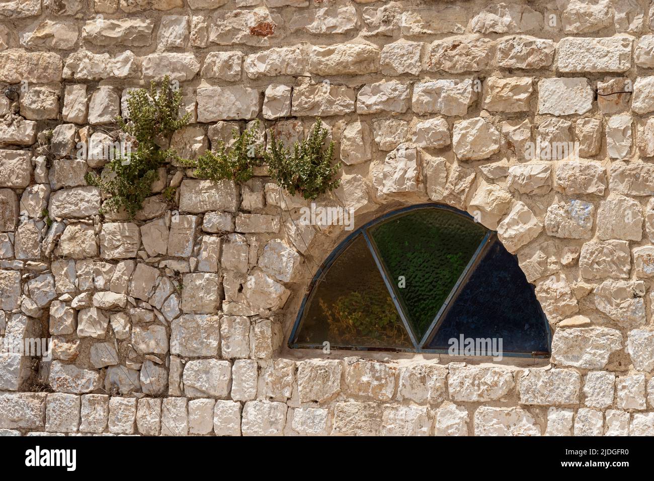 plants grow in cracks in an ancient limestone wall in Safed Tsfat in Israel next to a half circle window with colored glass triangles Stock Photo