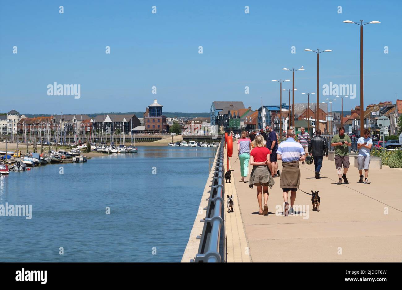 Littlehampton, West Sussex, UK. People walk along the riverside fottpath on the East bank of the River Arun. Shows town centre and yachts beyond. Stock Photo
