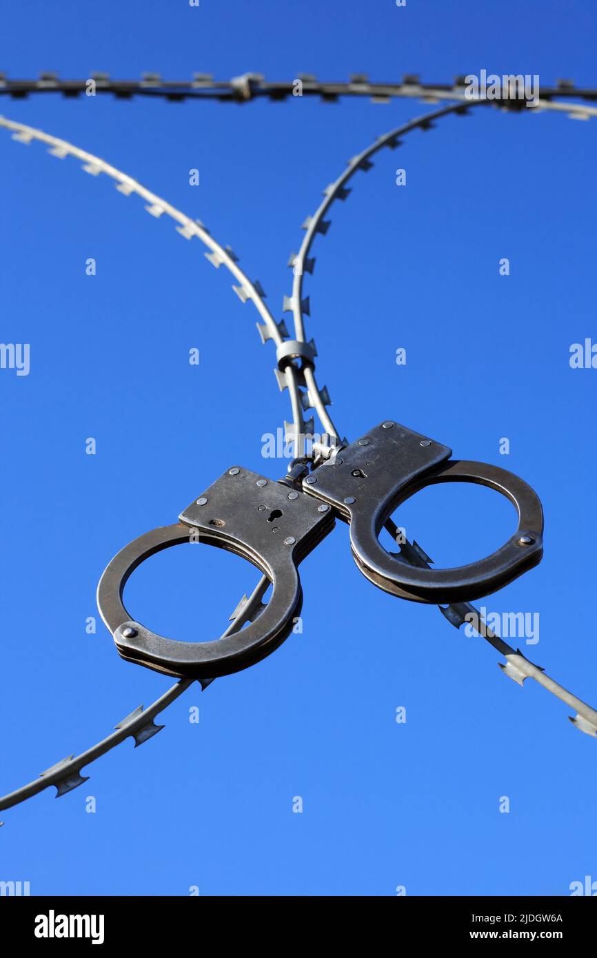 Old metal handcuffs hanging on razor wire against blue sky Stock Photo