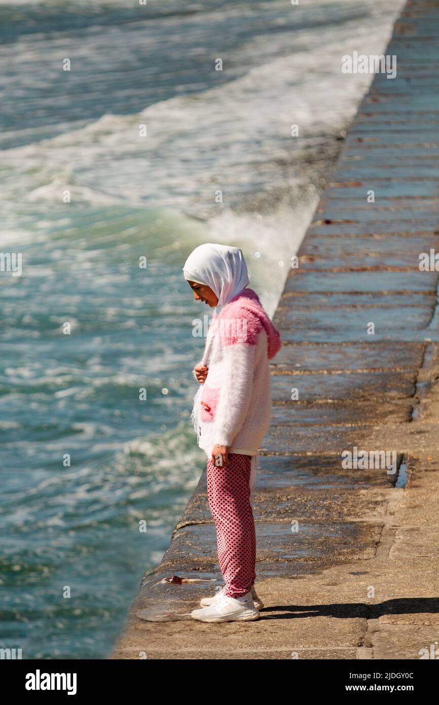 Female Muslim child refugee watches the surf break against the promenade Stock Photo