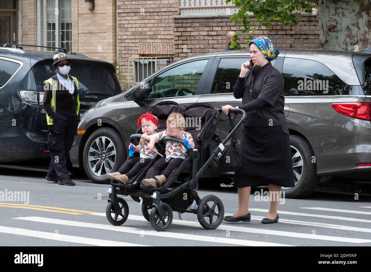 An orthodox Jewish woman pushes her children in a stroller while chatting on her cell phone & under the watchful eye of a crossing guard. In Brooklyn. Stock Photo
