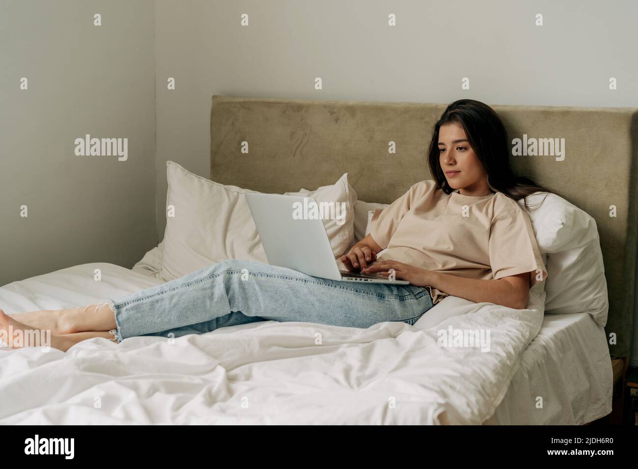Young brunette freelancer in t-shirt and jeans lying on bed working on laptop Stock Photo