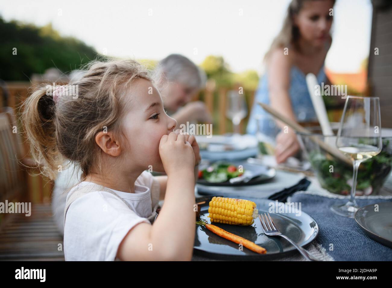Family eating at barbecue party dinner on patio, little girl eating roasted corn and enjoying it. Stock Photo