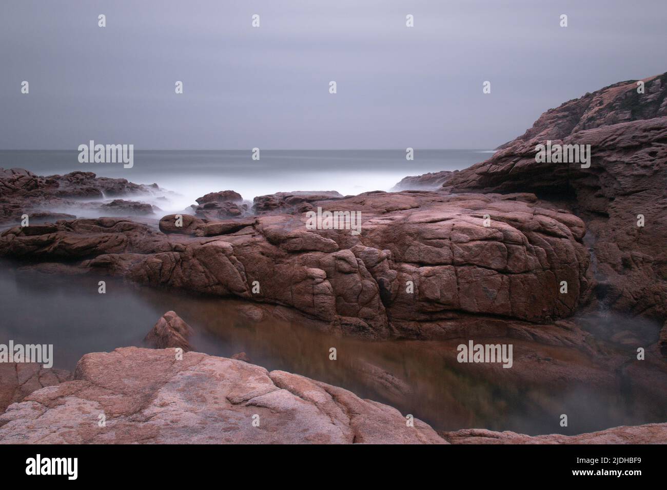 Very long exposure of waves breaking on rocks.  Dull grey sky overhead. Stock Photo