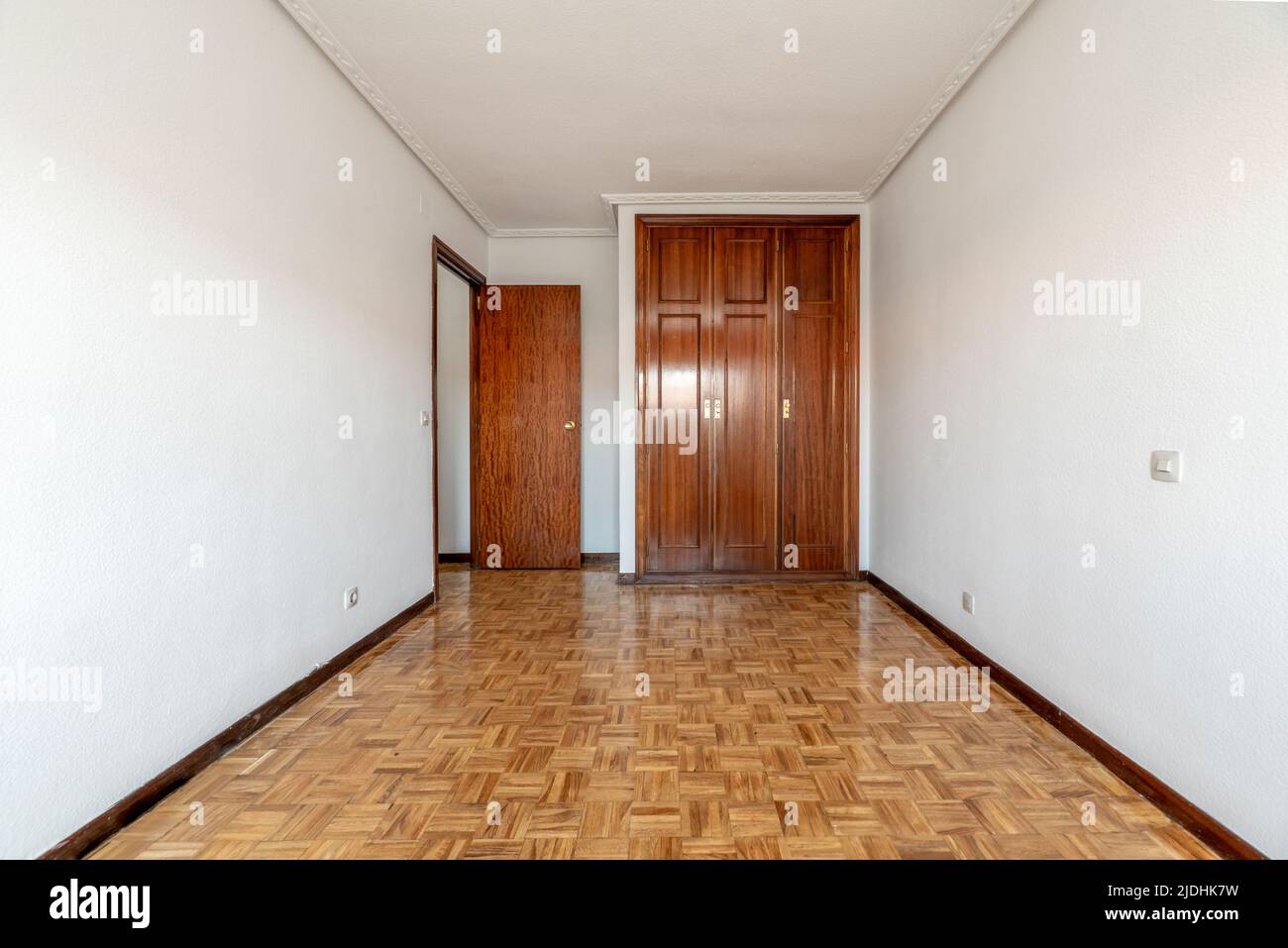 Empty room with shiny oak parquet flooring, plain white painted walls and reddish woodwork on doors and built-in wardrobe Stock Photo