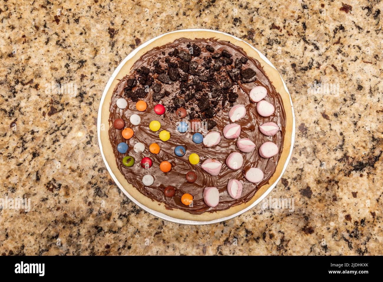 Sweet pizza with nutella, pieces of chocolate biscuit, marshmallows, assorted chocolate sprinkles on granite table Stock Photo