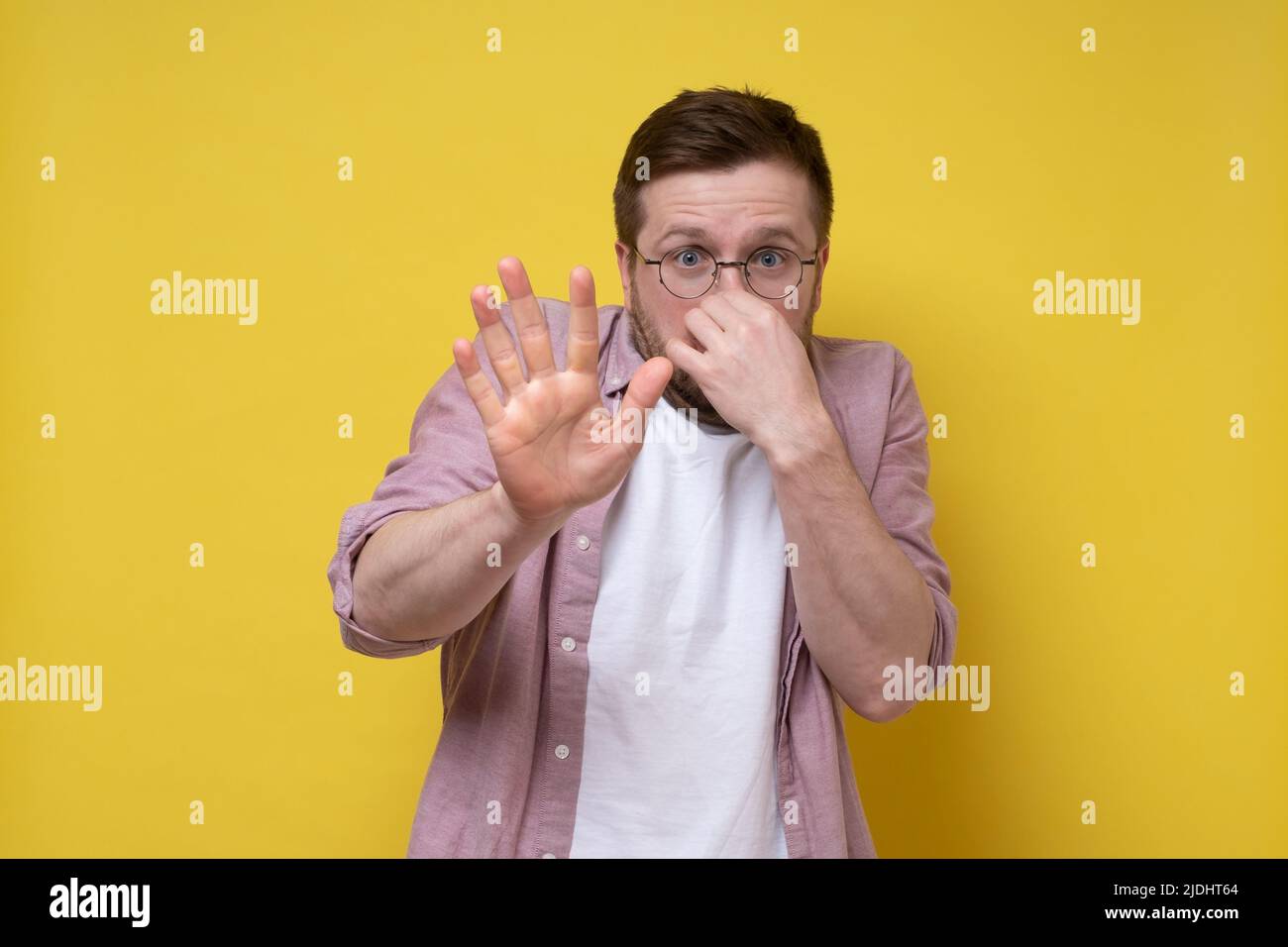 Young man smells an unpleasant smell, he pinches nose with fingers and makes a stop gesture with hand. Yellow background. Stock Photo