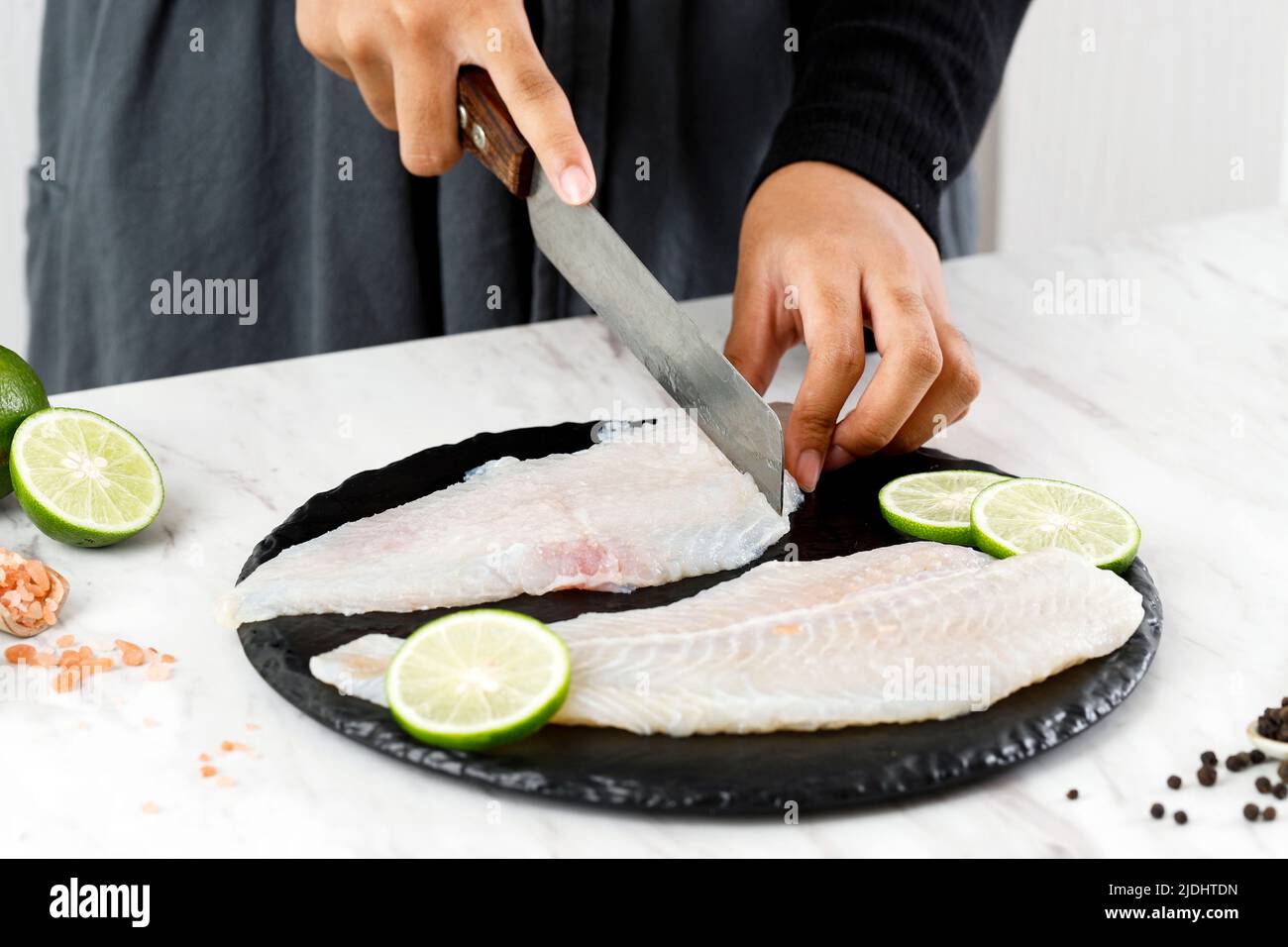 Female Hand Cutting Dori Fillet Pangasius Fish above Stone Board, Process Cooking in the Kitchen Stock Photo