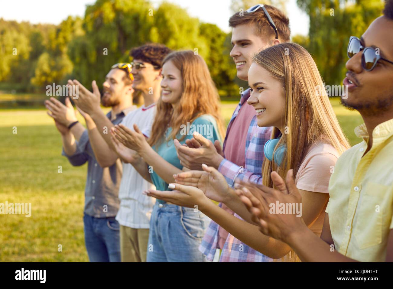 Group of joyful contented multiracial young people applaud and cheer together in park. Stock Photo