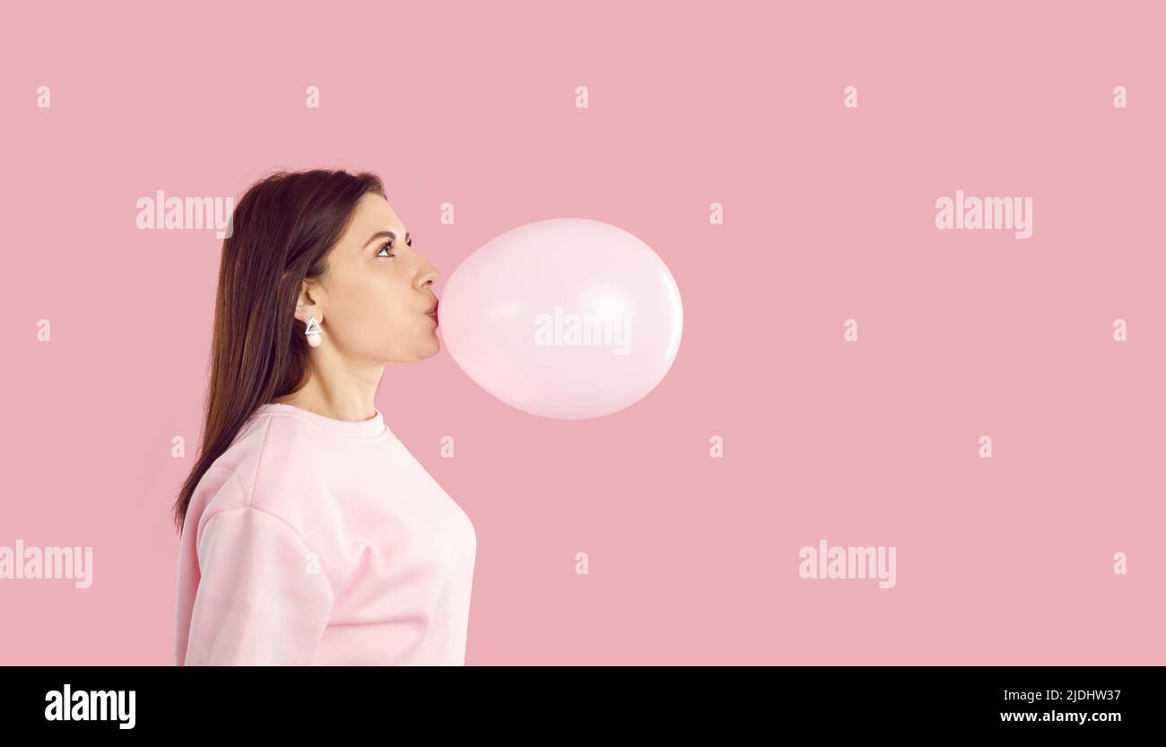 Side view of happy young woman blowing up pink balloon on pink copy space background Stock Photo