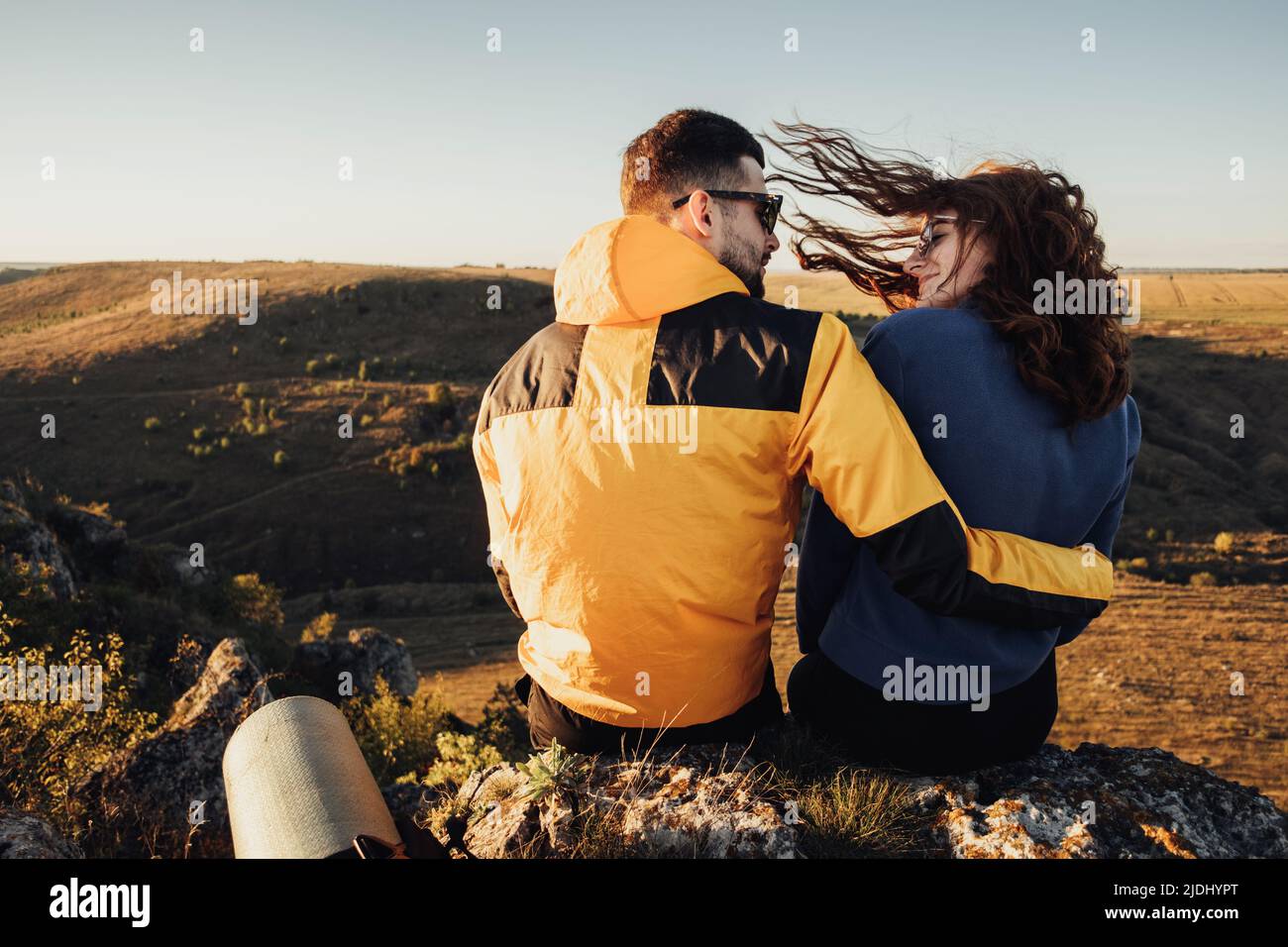 Back View of Traveler Couple Hugging and Sitting on the Edge of Hill During Sunset, Wind Blowing Woman's Hair Stock Photo