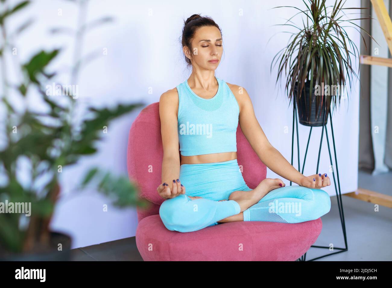 A woman in blue sportswear practices yoga, sits in a lotus position in a chair and meditates with her eyes closed Stock Photo