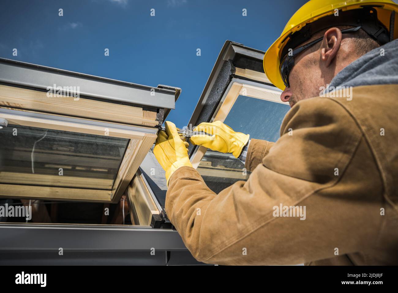 Caucasian Worker Wearing Safety Helmet and Protective Gloves Carrying Out Repair Works on Roof Skylight Windows Using His Screwdriver. Stock Photo