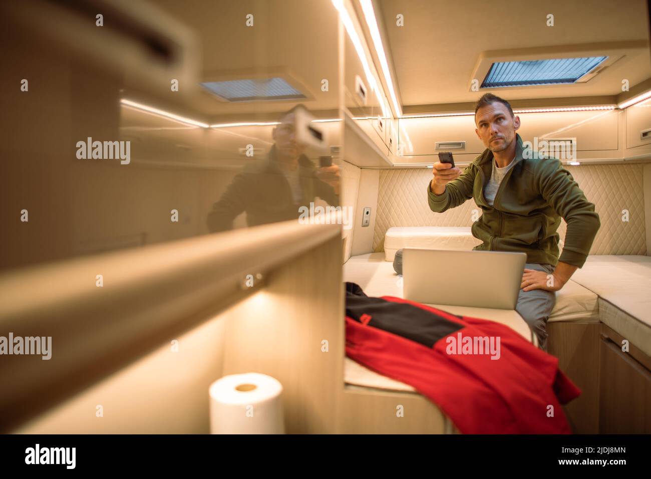 Caucasian Male Traveler Switching On the TV with Remote Control While Resting in the Sleeping Zone of His Recreational Vehicle During the Road. Stock Photo