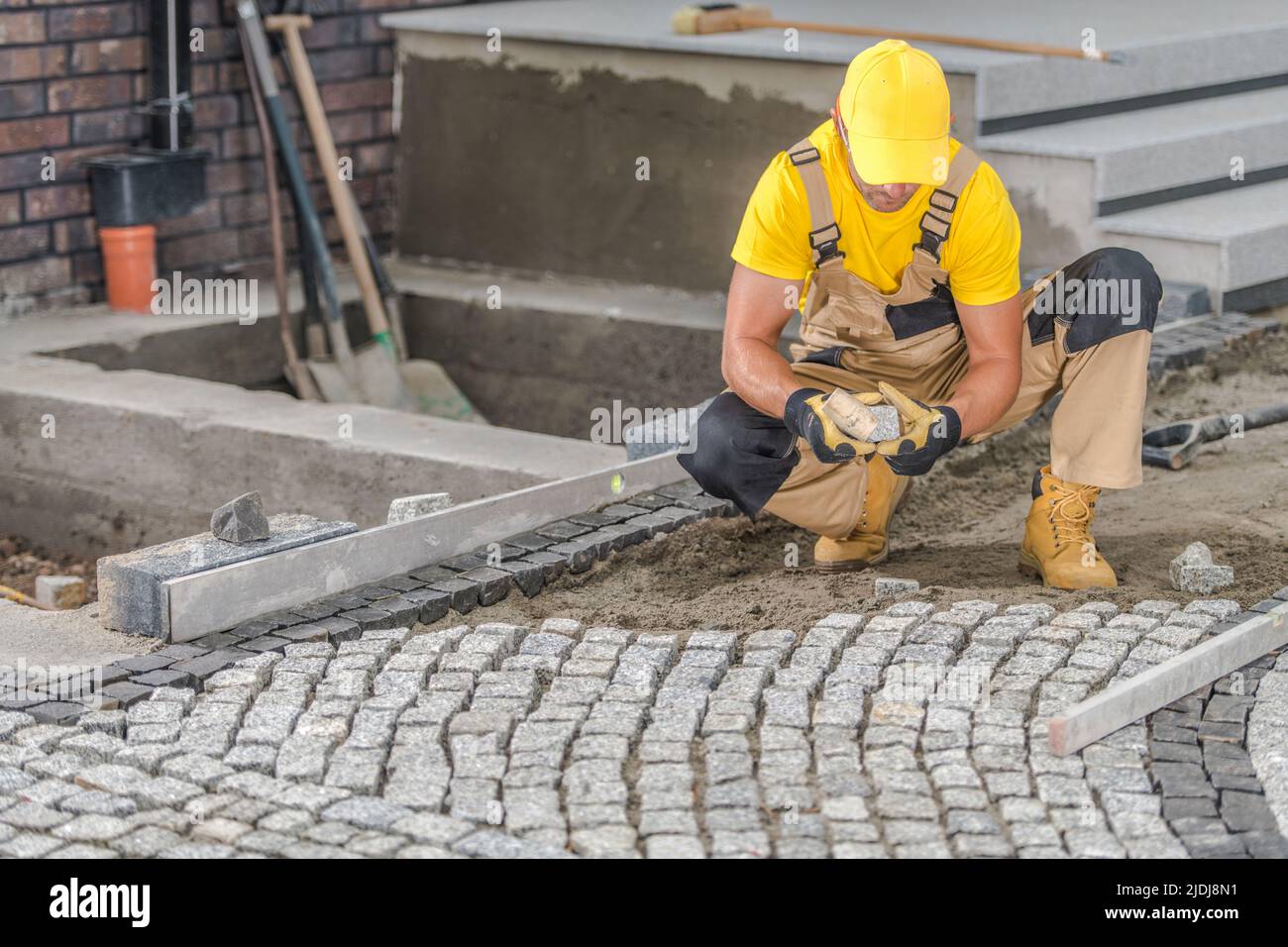 Caucasian Contractor in His Work Wear Carefully Laying Granite Setts in His Client's Front Yard. Landscaping of the Area Around a Newly Constructed Re Stock Photo