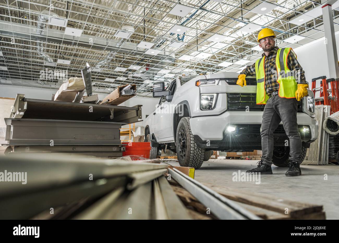 Caucasian Worker Wearing Yellow Hard Hat Inside Developed Warehouse Building Staying Next to His Modern Pickup Truck Commercial Vehicle. Stock Photo
