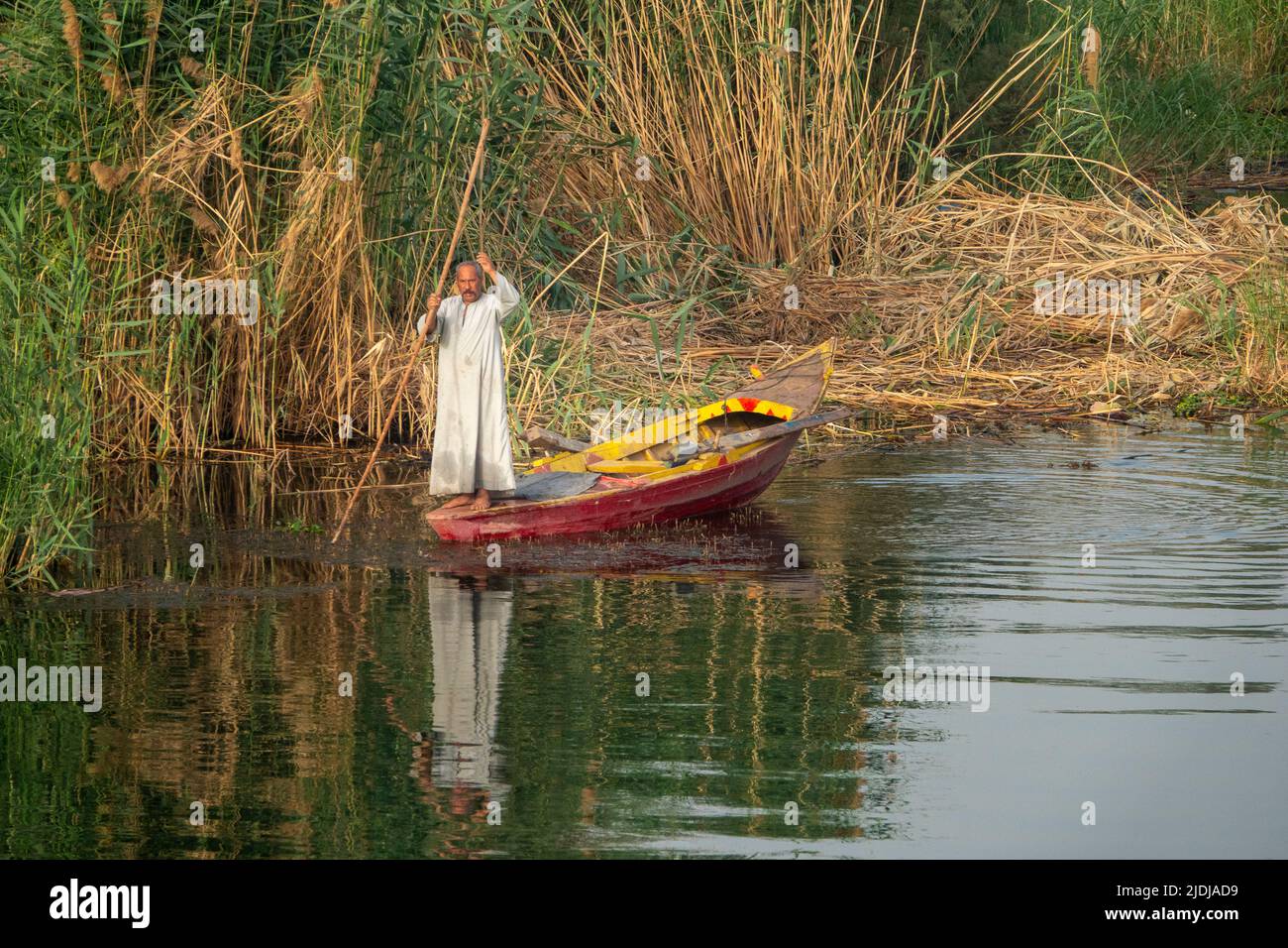 A lone Nile fisherman holding long stick standing in brightly painted wooden fishing boat in a small creek with gently rippling water and reflections Stock Photo