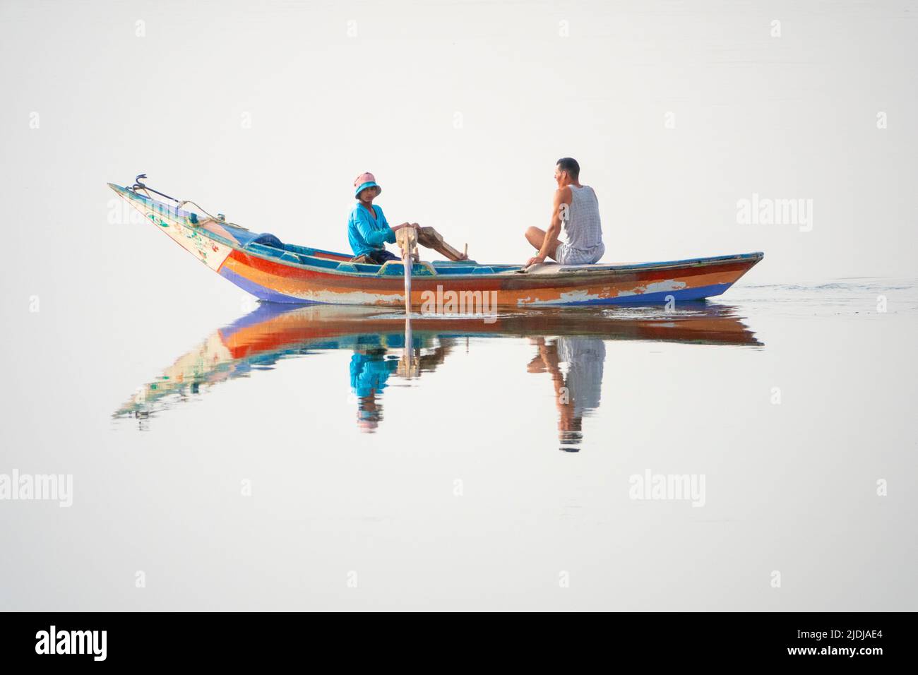 Two young men, one rowing and one on mobile phone in a brightly painted wooden boat perfectly reflected in mirror surface water Stock Photo