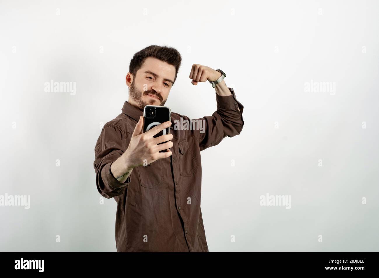 Caucasian man wearing brown shirt posing isolated over white background taking selfie, showing strong arm, flexing biceps and posing for photo. Stock Photo